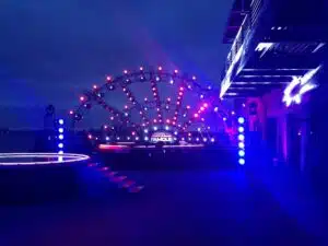 Half circle truss arches over a round stage in a half-shell configuration. It is night. there is a blue wash over the area with redish-pink lights along the truss arches. The sign in the very back of the stage says "Make a Band Famous".