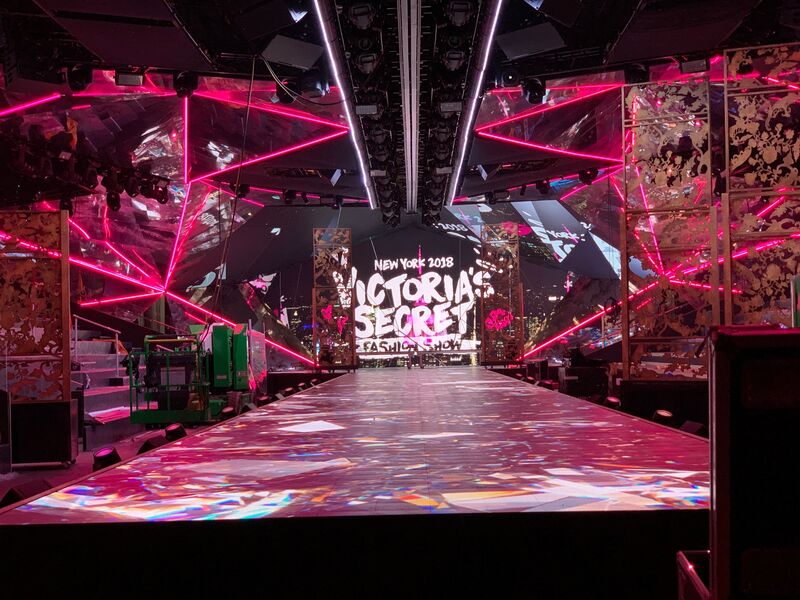 Victoria's Secret stage, looking down center of stage from audience POV. Stage is washed in pink and white with the Victoria's Secret logo on a video wall in the center upstage.