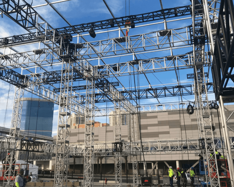 Workers work under a large, multi-layered outdoor truss structure.