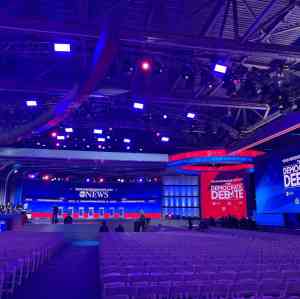 Production rigging for the 2020 democratic debate for ABC news. Lots of truss and lights are rigged from a venue roof. Rows of white chairs under a blue lights are in front of the set. United Staging & Rigging