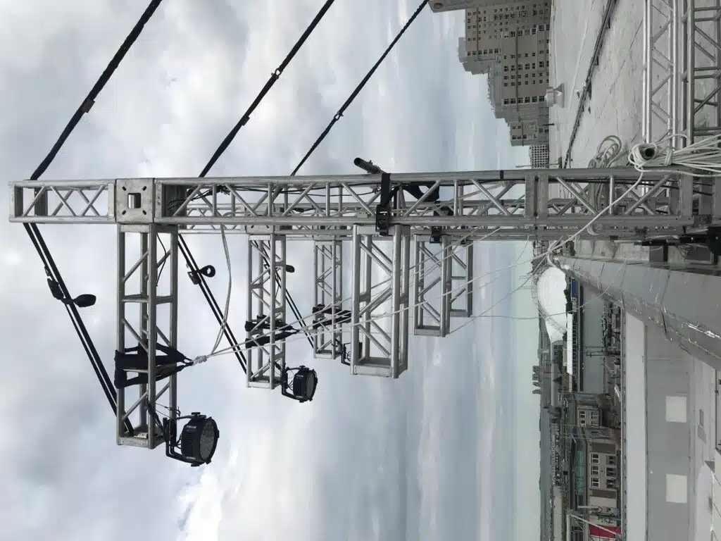 Truss towers outside on a roof with cantilever truss hanging over edge of roof, to hang lights from the end of each cantilever, Lights point down towards ground. You can see safety lines pulling back out of the frame to hold the trusses in position.