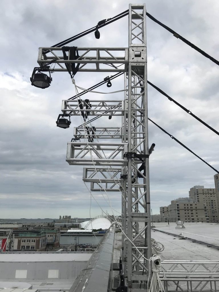 Truss towers outside on a roof with cantilever truss hanging over edge of roof, to hang lights from the end of each cantilever, Lights point down towards ground. You can see safety lines pulling back out of the frame to hold the trusses in position.