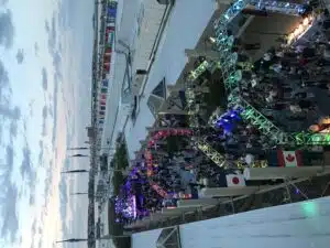 Five angular truss arches in a row, all lit up, during an outdoor event with people milling around underneath of them. A canada and japan flag hang (there are more but you can't make them out). Event is taking place on a roof of a building in the evening. POV is from above the event looking down onto it