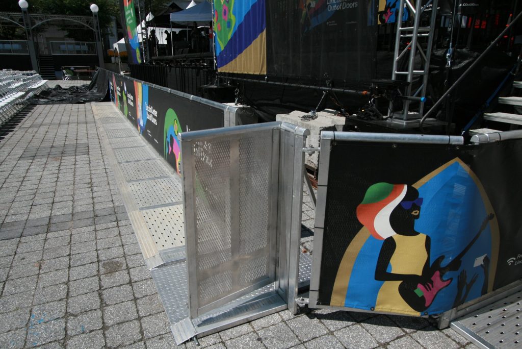 Line of silver barricade on ground of gray pavers. A barricade door is closest to the camera and is open. the rest of the barricade is covered in black with bright colors festival banners. There is a stage behind this you can only see the bottom half of.