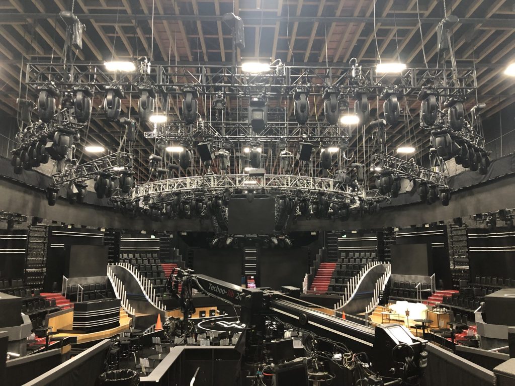 chain motors hanging a circular grid with lights and some straight sticks of truss, also with lights, surround the circular truss. a camera jib is below the trusses on the ground. inside of a filming area.