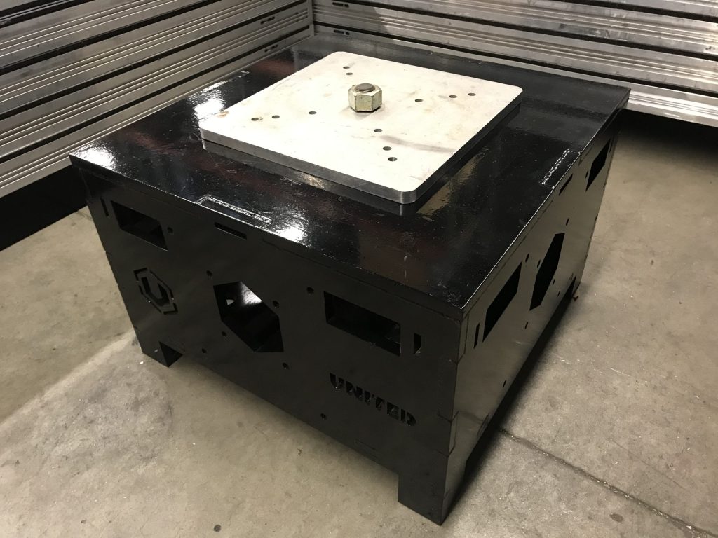 United Staging & Rigging's patented UNI-Bloc stage ballast. Looking down at the top and corner. Its a black cube with a silver top adjustable plate that can be tightened into place with a large bolt (shown). Sides have multiple holes for picking with forklifts, pallet jacks, or to access the interior of to add weight or store equipment. UniBloc sits on a warehouse floor.