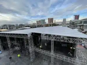 Huge outdoor truss structure on a partially cloudy day. A gray keder roof skin covers the whole thing. Large city buildings are in the background. POV is above the roof looking down. A few people are on the ground under the structure. United Staging & Rigging
