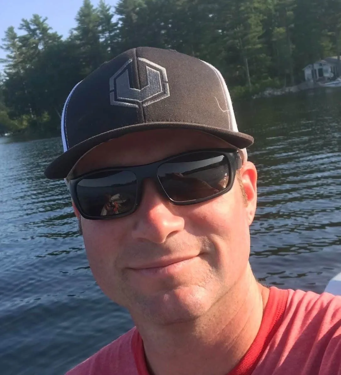 Face of man in red shirt, sunglasses, and black hat with the United Staging "U" logo on it. He is in front of water and trees, from the angle it looks like he's on a boat