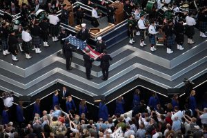 Stage risers in a zig zag pattern with a crowd below it. A line of people in blue walk in the space between the crowd and the base of the risers. On the upper right and left sections of the riser are band members in kiltz. In the center of the risers are military men in uniform folding an American flag. This is a ceremony. POV is looking down onto the risers. United Staging & Rigging