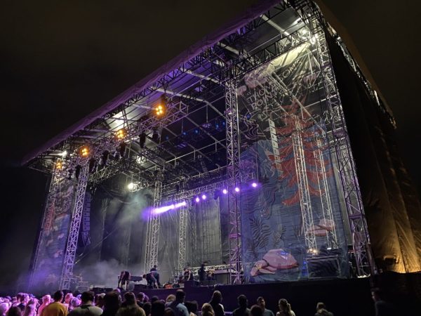 POV: Your in the crowd, at night, towards the right side of an outdoor concert stage with purple lights washing the stage. United Staging & Rigging