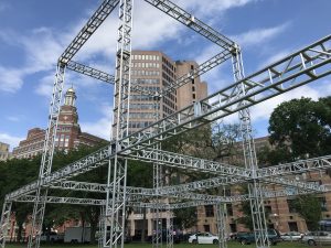 A large multi-cubical truss structure outdoors. on a mostly sunny day, in front of brown, brick buildings. Cars line the streets behind the structure. United Staging & Rigging