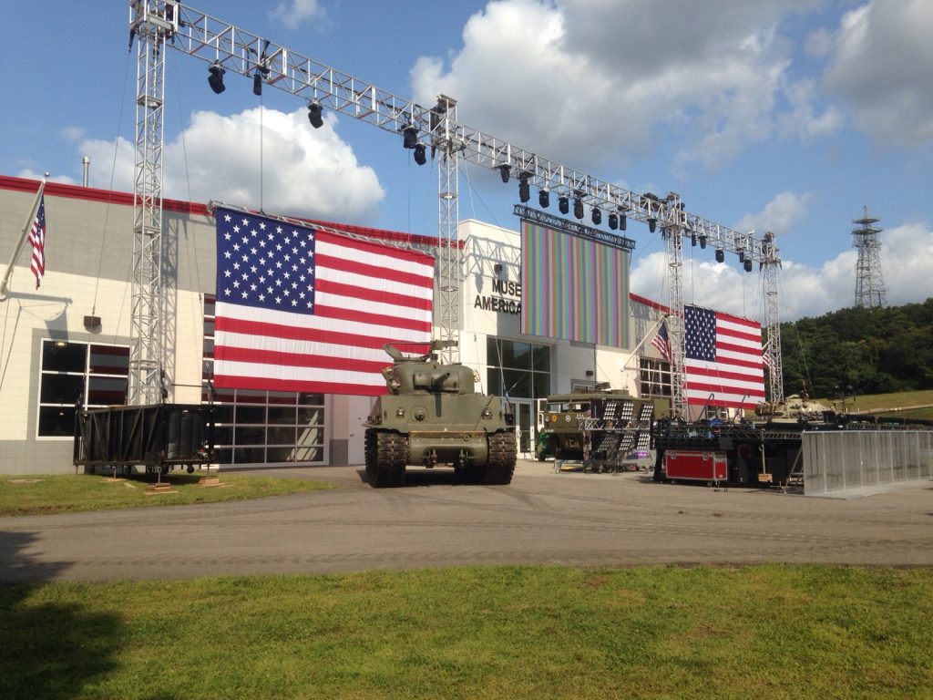 Three connected rectangular truss arches with lights and American flags hanging from it, in front of a building. You can see a partial sign for the building in the background ("Muse" & "America") An LED screen hangs from the center arch. An green army tank is on the ground below the nearest truss arch. This was from the set of an Impractical Jokers episode.