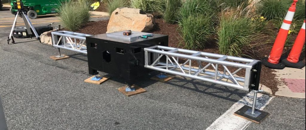 Uni-Bloc, United Staging & Rigging's patented stage ballast on the ground outside with a short stick of 12" truss attached to opposite sides. It's sunny out and tufts of landscaped grasses are behind along with safety traffic cones.