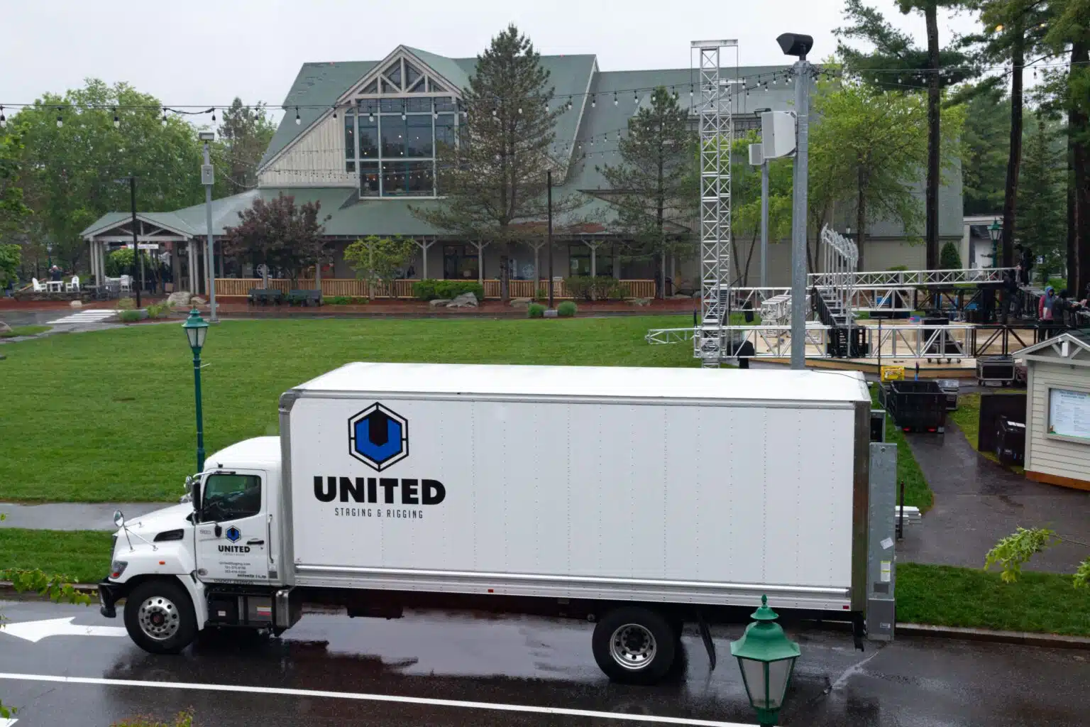 White medium size box truck with blue "United Staging & Rigging" logo on the side and door. POV is side of truck from above (photo taken out of the second story window of LLBean's Hunting shop in Freeport Maine). Truck parked on pavement with grass behind it.
