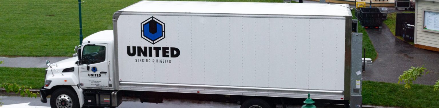 White medium size box truck with blue "United Staging & Rigging" logo on the side and door. POV is side of truck from above (photo taken out of the second story window of LLBean's Hunting shop in Freeport Maine). Truck parked on pavement with grass behind it.