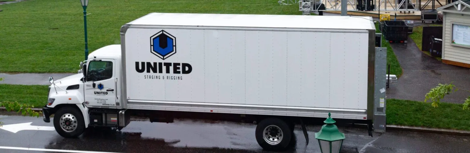 White medium size box truck with blue "United Staging & Rigging" logo on the side and door. POV is side of truck from above (photo taken out of the second story window of LLBean's Hunting shop in Freeport Maine). Truck parked on pavement with grass behind it.