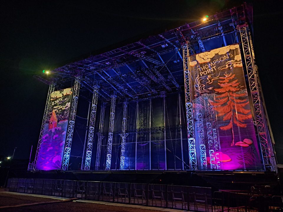 Outdoor stage at night with blue lights and "Thompson's Point" and "State Theater"banners on the front. POV is from the audience right of the stage. You can see up into the underside of the roof part to see all the trusses, towers, and staging and rigging structure. The sky is black. Barricades are on the ground in front of the stage. United Staging & Rigging