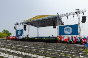 3/4 view of an outside stage with a yellow roof and two blue logo'd school banners hanging from the wings on either side of the stage. Flags from all countries decorate the front of the stage wings and rows of metal, folding chairs are in front of the stage. The stage is large. The roof structure is 45 ft by 65 ft.