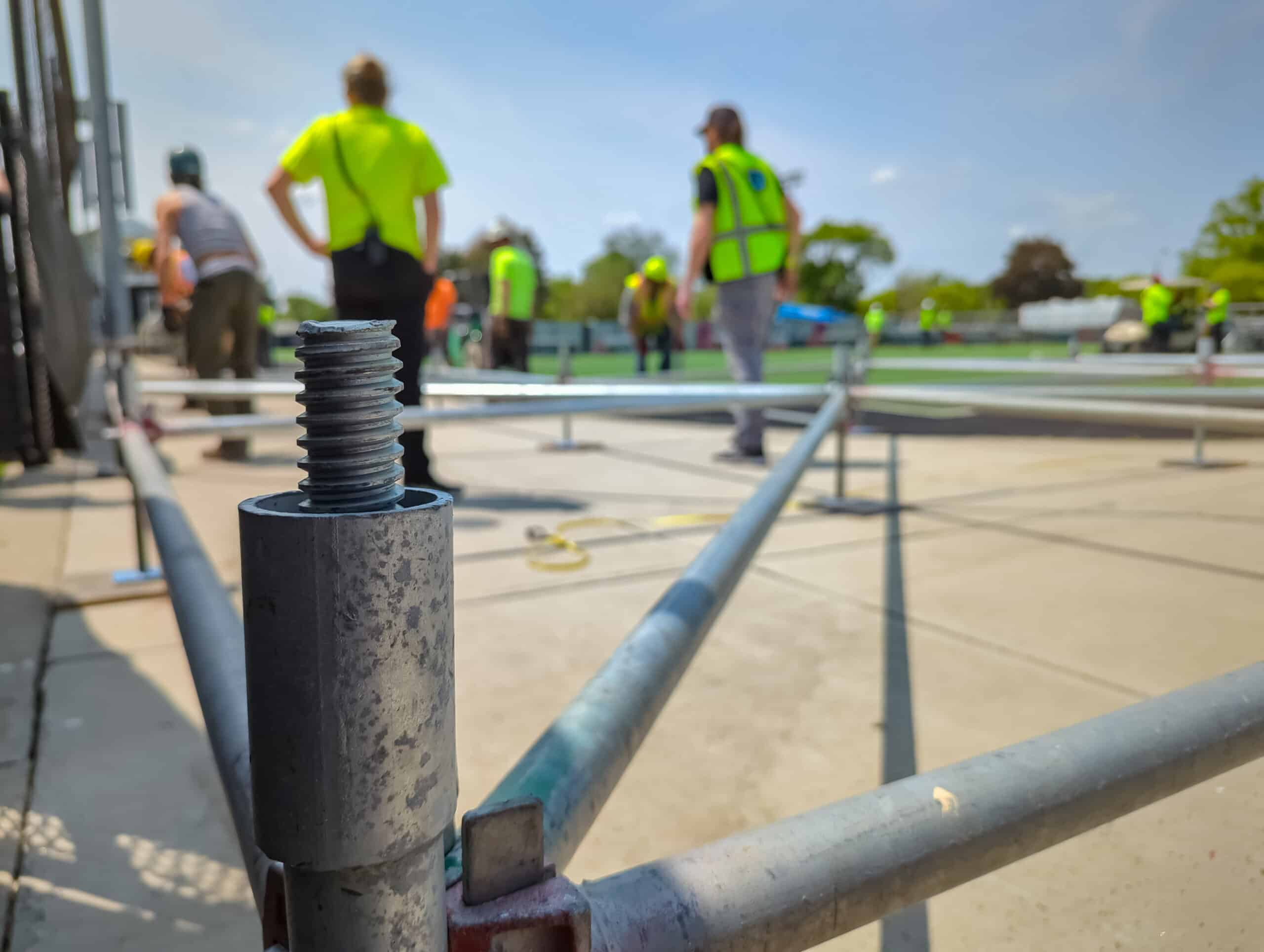 Cropped view of a scaffold build being laid out, with a steel piece in focus in the front and some layour with unidentifiable crew members' legs in the background.