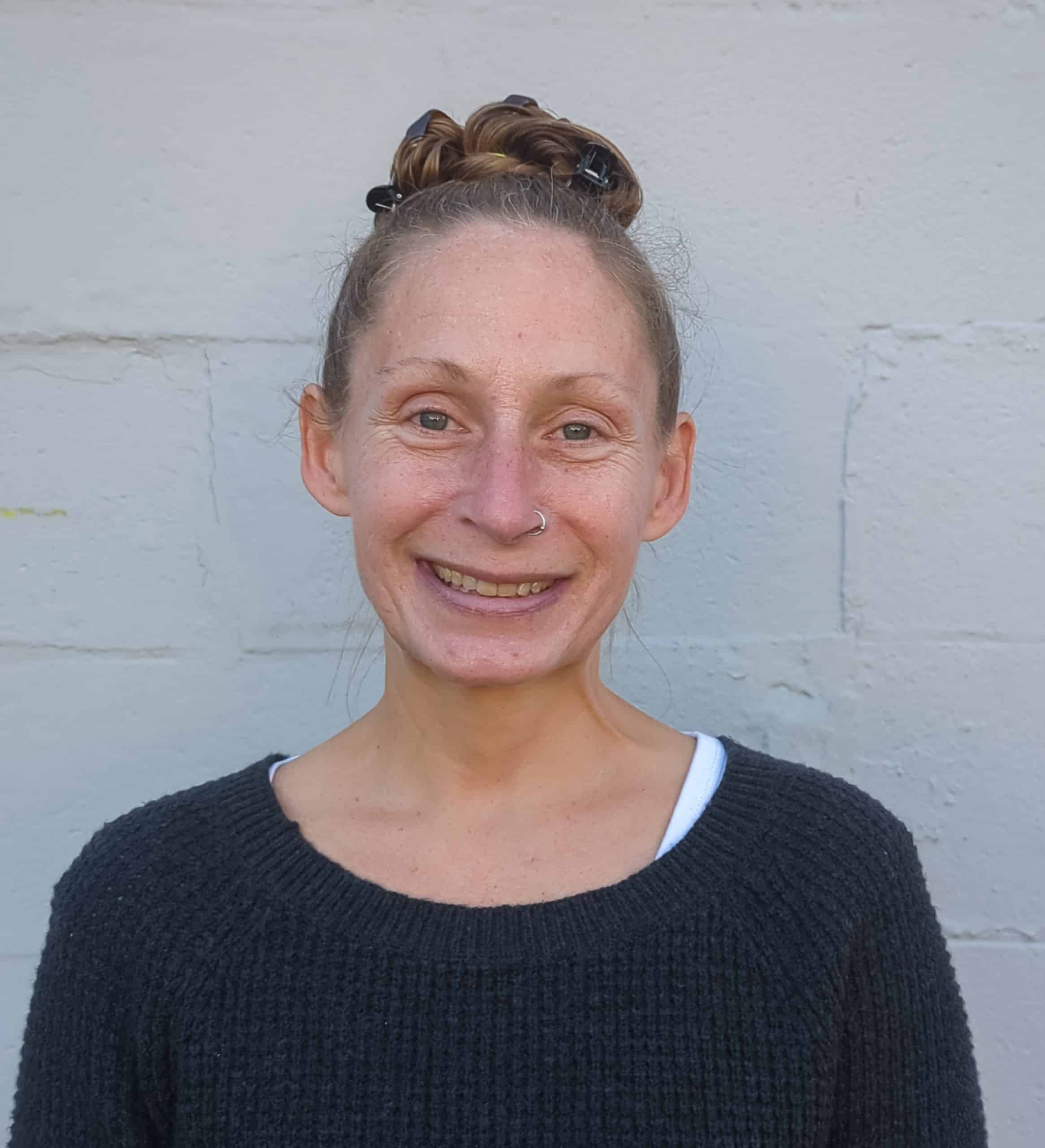 Chest up photo of a smiling woman standing in front of a white painted brick wall. Her light brown / dirty blonde hair is in a bun on the top of her head and she wears a black sweater with a white tshirt peeking out from underneath.