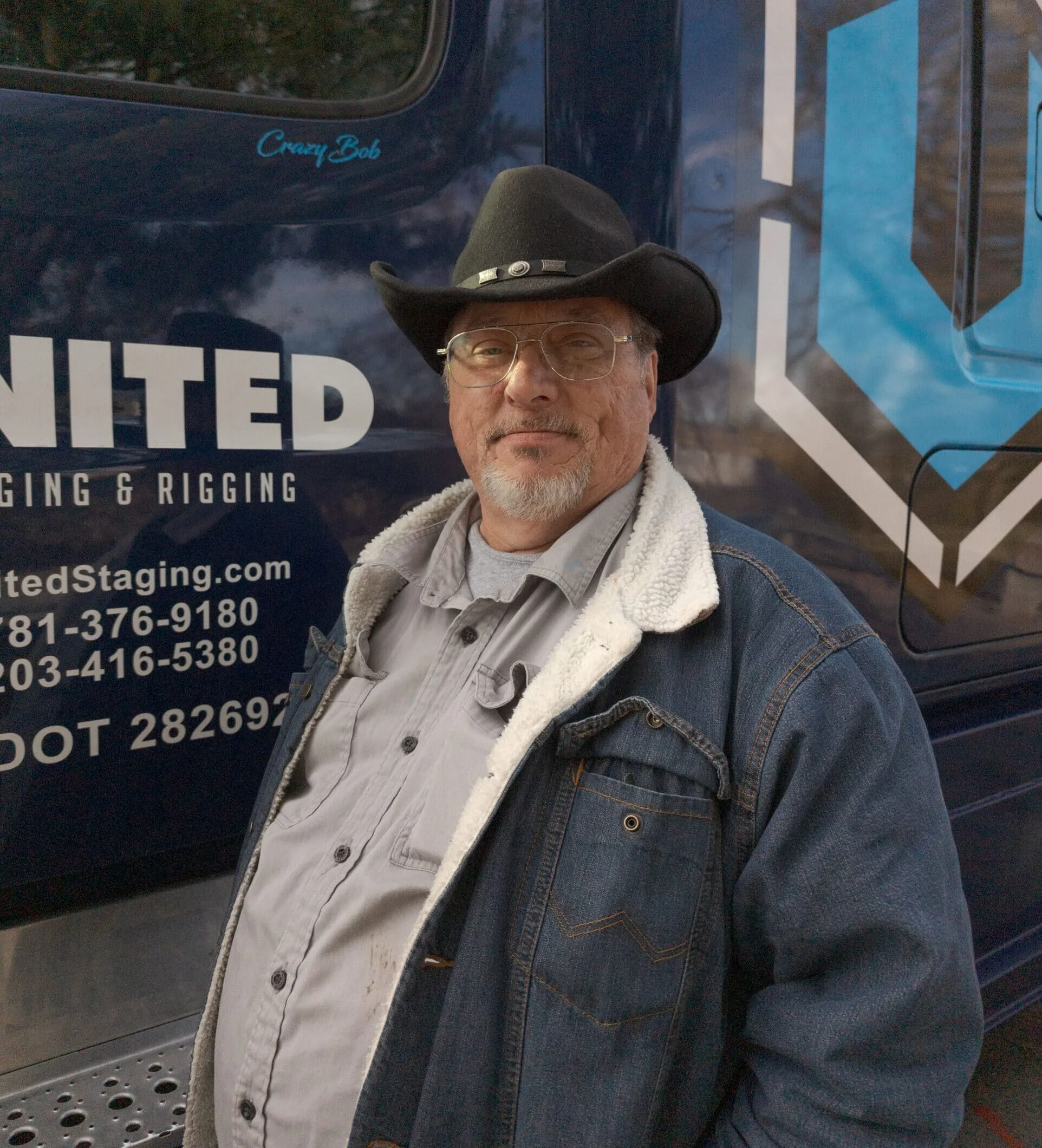 Man in dark blue jacket and black cowboy hat standing in front of the dark blue cab of his semi truck