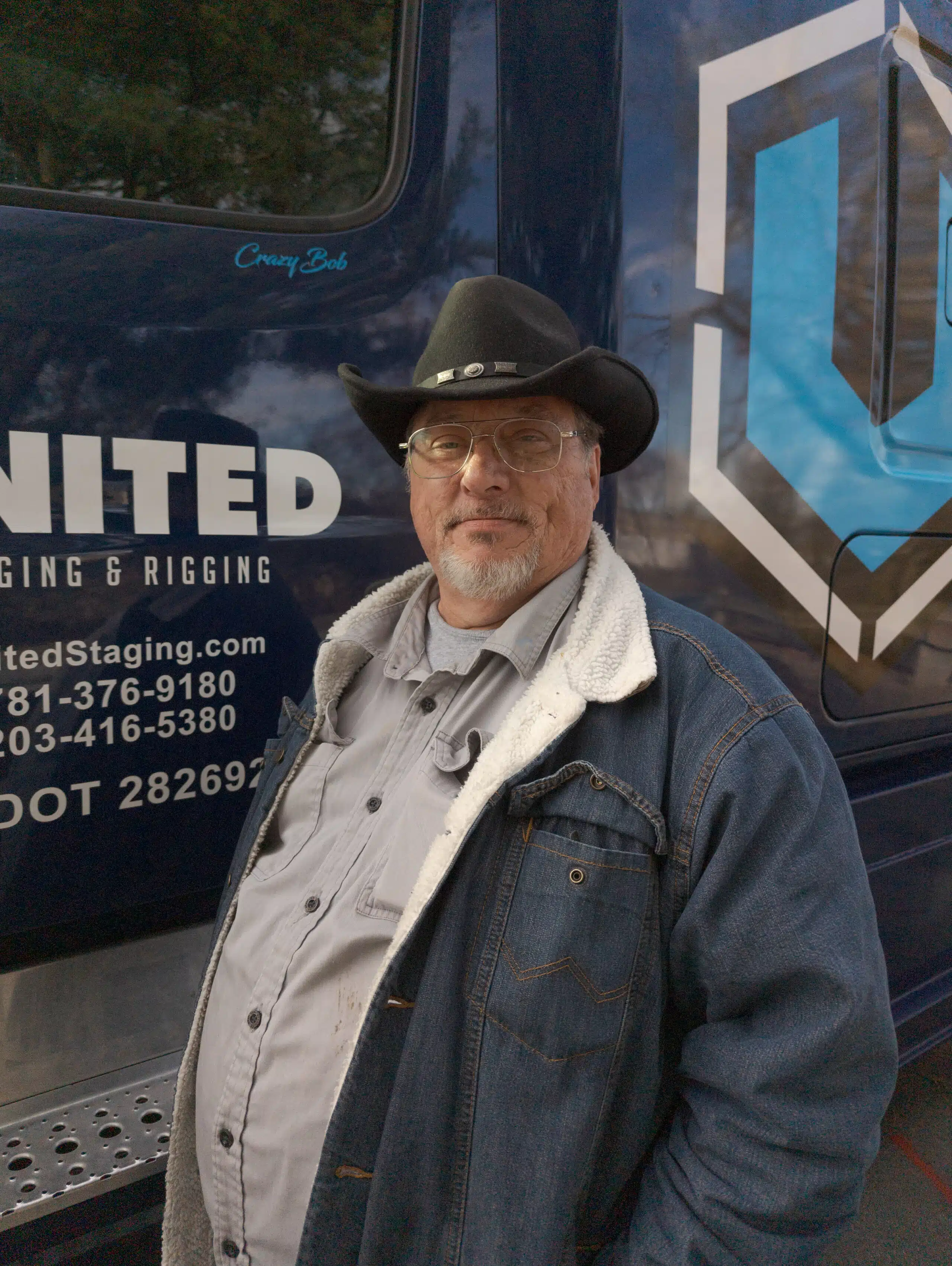 Man in dark blue jacket and black cowboy hat standing in front of the dark blue cab of his semi truck