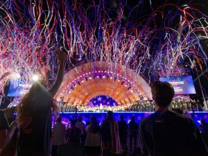 red, white, and blue streamers fly overhead of the audience during the 4th of july show at the hatch shell in boston. POV: You are in the crowd a few rows back from the front looking up, with the silhouettes of a man and girl in fromt of you jumping in the air excited for the streamers. The stage is mostly light up in blue at this point.