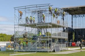 Climbers on multiple levels of a 3 story scaff structure are building what will be front off house for Boston Calling 2025. The workers all wear hard hats and safety yellow or orange shirts and vests. the structure doesnt have any wall or roof skins on it yet but there are handrails along the sides and back edge for protections. some climbers are poisitoned on the verytop in harnesses getting ready for a person on the bottom right ground to start sending up the grey roof skins (also laid out on the ground).