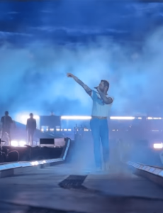 Post Malone walking down the centerramp during his "big ass stadium tour". Everything is washed in blue light and haze. Post holds the mic with one hand and raises his other pointing out into the crowd. View is FPV from right in front of the end center of the ramp with no one in front of them. This is a screenshot from a video of the show.