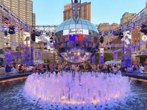 A giant disco ball hangs over a central water fountain. The fountain water in the basin is blue and the circle of water spraying up slightly is lit up in pink. Overhead you can see some of the custom truss structure that holds up the disco ball and various lights and other decorative elements. Thsi whole thing is outdoors during the day (looks like morning low light), and sits in JR Plaza at Lincoln Center.