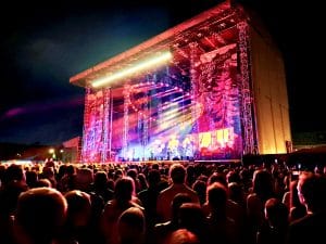 A large outdoor stage in the black of night is lite up in pinks and yellows. A round lighting truss sits at an angle so the lights face the crowd diagonally and light shoots from the lamps onto the crowd below. Standing to the right of the stage you can see the crowd and full stage aglow and feel the energy of the evening.
