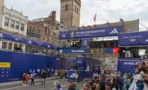 The boston marathon frinishing line for the 5k race that happens the day before the marathon. many dark blue signs cover the giant structure that's built across the stree and crowds are gathered on the right side in the stands. (the left side is just a blue backdrop). Some runners and event team members are in the actual street. The signs say 129th Boston marathon, bank of america (a sponsor) and WCVB5 (tv station). The lower of the overstreet signs has a countdown clock on an LED screen. The screen says ProWomen on the left and the clock has 16:45.0. Its a partly cloudy morning. Olf typical boston brick buildings and a church tower are in the background.