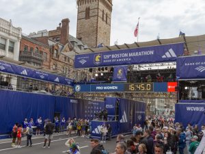 The boston marathon frinishing line for the 5k race that happens the day before the marathon. many dark blue signs cover the giant structure that's built across the stree and crowds are gathered on the right side in the stands. (the left side is just a blue backdrop). Some runners and event team members are in the actual street. The signs say 129th Boston marathon, bank of america (a sponsor) and WCVB5 (tv station). The lower of the overstreet signs has a countdown clock on an LED screen. The screen says ProWomen on the left and the clock has 16:45.0. Its a partly cloudy morning. Olf typical boston brick buildings and a church tower are in the background.