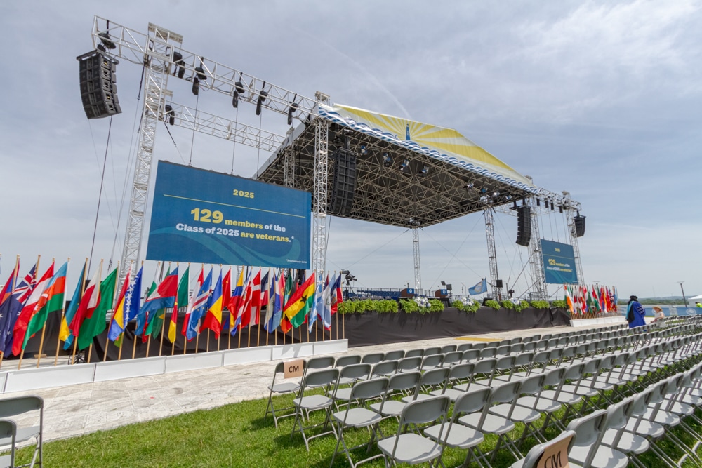 VIew from the audience left side of the UMASS boston commencement stage on show day. The decorations have been put up. There is a stage with a roof graphic that is a a lighthouse in the water with yellow light beams emminatig from the lighthouse. the front of the stage here is adorned with green foliage and the base is covered with black fabric (to hide the understructure of the stage). On each side of this main stage are stage wings: open truss structures that hold speakers and video walls. The video walls both have a blue background with the words 2025, 129 members of the class of 2025 are veterans. Flags from all different countries blow in the wind under these side wings. The photographer is standing in the left center audience aisle a few rows back. you see grass on the ground and some rows of grey chairs to the side. The sky is a light blue with whispy white clouds and its indirectly sunny (you dont see the actual sun).