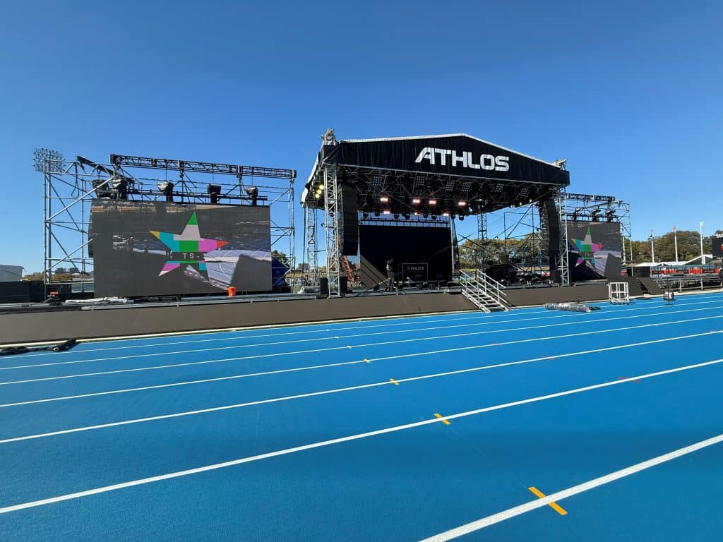 Stage with side video wall wings. Stage has black header that says "ATHLOS" in white letters. In the foreground, in front of the stage, is a medium blue running track.