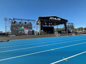 Stage with side video wall wings. Stage has black header that says "ATHLOS" in white letters. In the foreground, in front of the stage, is a medium blue running track.