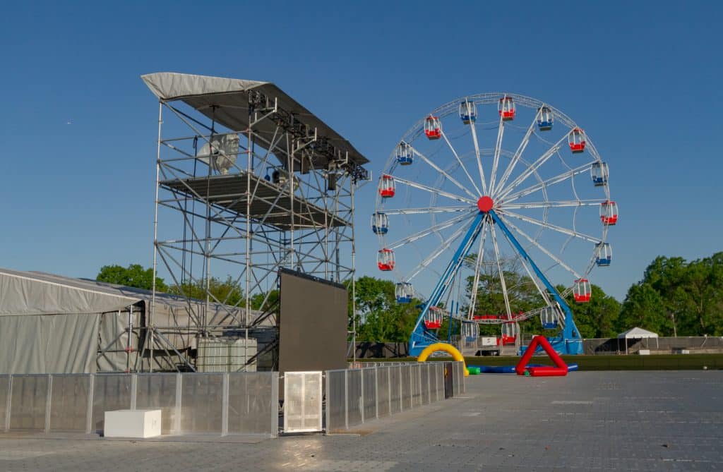 Crowd control barricades block off a scaffold foh structure on a festival ground. A ferris wheel is in the background