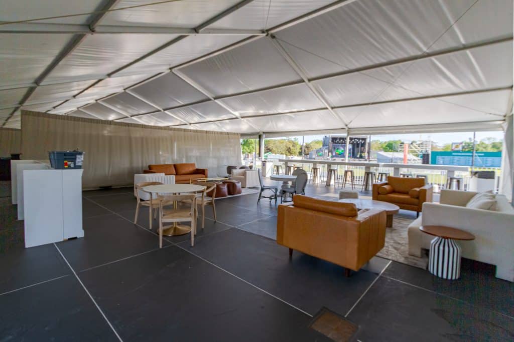 inside the VIP area at boston calling festival. The floor is made of stage decks, eleveated above the ground on scaff structure. white roof and, side, and backwalls protect VIPs from the elements, while the open front and elevated height give them an unobstructed view of the concert stages. The area is decorated with rust-orange, white, and light-colored wood chairs, couches, and side tables arranged in livingroom style, with big open middle spaces - in a way that encourages conversation.