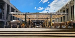Truss Structure outside with open-air roof on which hangs many rows of golden-beige fabric strips that move in the wind. Underneath this stucture is a plaza decorated in golden decor for Lincoln Center's 2026 Summer For the City event season.