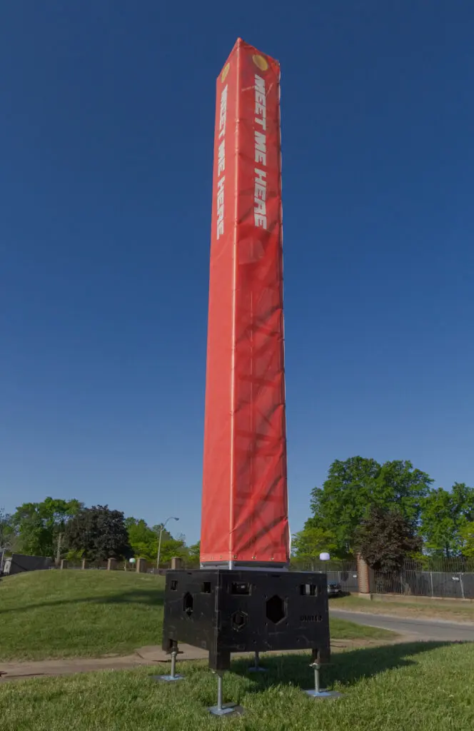 Red festival sign rises vertically into the air ontop of a UNI-Bloc ballast thats in a green grassy field.