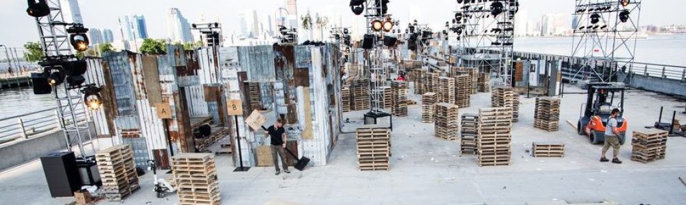 Lot of truss towers with lights amongst stacks of wooden pallets and other scenic pieces on a barge or wharf on the water. a few workers are visible.