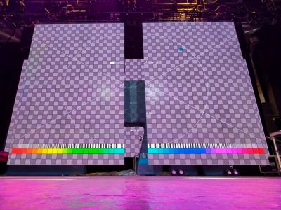 Pink and lavendar lighting cast over a stage an automated LED video wall thats partially open. The video wall has a checkerboard test screen image on it and a color bar test strip projected at the bottom. The wall is in the process of opening and you can make out the automation truss through the darkness at the top of the wall. Your view is from a low angle on the stage.