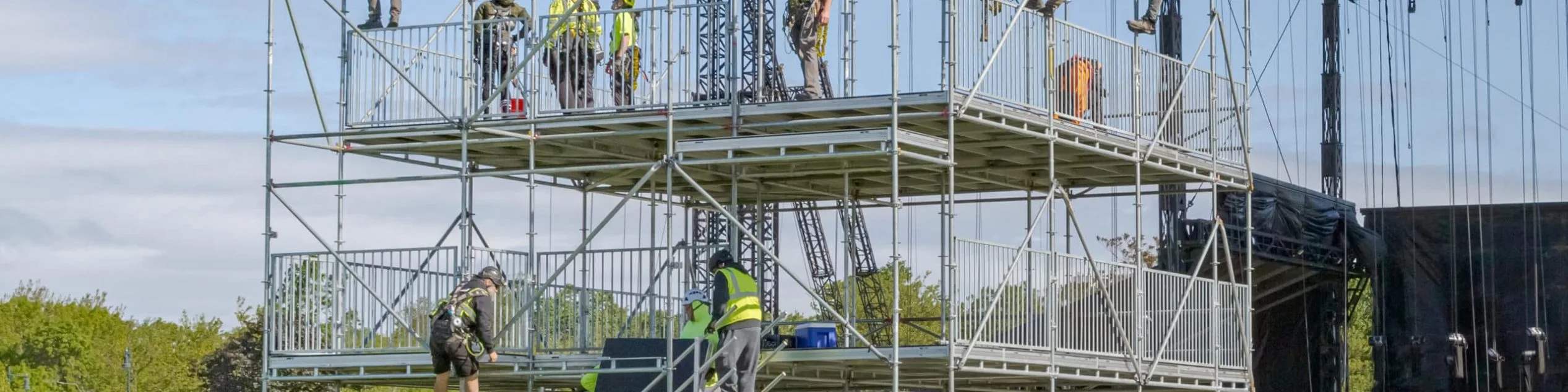 Riggers and stagehands on various levels building a three-story scaff structure that will be the front of house tech area for Boston calling 2025. On the top level, riggers in harnesses are hanging from the top of the structure getting ready to add vinyl skins to create a waterproof roof. A couple site supervisors are on the ground instructing different teams on projects to complete this. Behind the structure, another company's stage is being set up. Everyone is outsie on a grassy field under a bright blue sky.