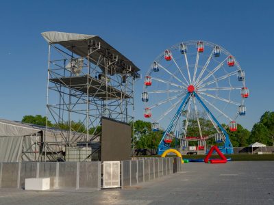Crowd control barricades block off a scaffold foh structure on a festival ground. A ferris wheel is in the background
