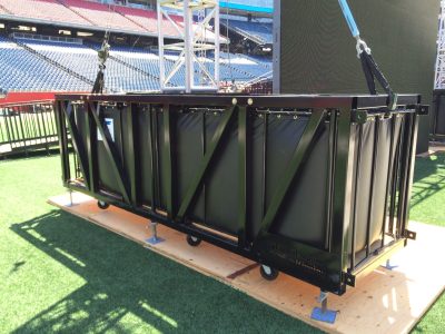 A large, black, rectangular steel container sits on plywood thats on a grass field inside a stadium. The container is made of steel bars, and the sides are not fully solid. Inside the container, you can see the sides of a thick, vinyl water ballast thatis full. This is in front of a wilver truss and turned-off video wall. You can see steel attaching to the ends of the ballast, and goign up in diagonal lines but where the steel cables are attached to at the top is cropped out.