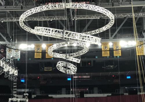 Multiple circular trusses hanging at various angles, with the diameter of each truss shrinking as the hang lower, creating a vortex or tornado shape. Circle trusses are hooked to automation motors in a mother truss "X" above it.