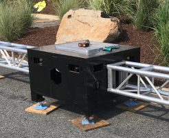 Close up of Uni-Bloc (United Staging's patented stage ballast) outside, on pavement, and in fron tof landscaped grasses. Silver truss are attached to opposing sides of the black Uni-Bloc. truss tools and a measuring tape sit on top of the adjustable top plate.