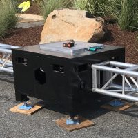 Close up of Uni-Bloc (United Staging's patented stage ballast) outside, on pavement, and in fron tof landscaped grasses. Silver truss are attached to opposing sides of the black Uni-Bloc. truss tools and a measuring tape sit on top of the adjustable top plate.