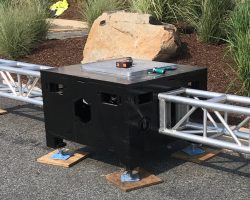 Close up of Uni-Bloc (United Staging's patented stage ballast) outside, on pavement, and in fron tof landscaped grasses. Silver truss are attached to opposing sides of the black Uni-Bloc. truss tools and a measuring tape sit on top of the adjustable top plate.