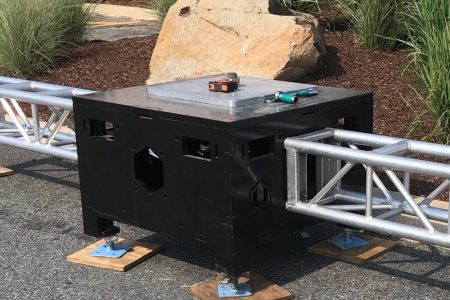 Close up of Uni-Bloc (United Staging's patented stage ballast) outside, on pavement, and in fron tof landscaped grasses. Silver truss are attached to opposing sides of the black Uni-Bloc. truss tools and a measuring tape sit on top of the adjustable top plate.